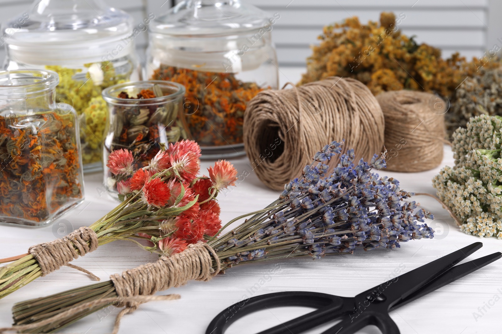 Photo of Bunches of dry flowers, different medicinal herbs, scissors and spools on white wooden table