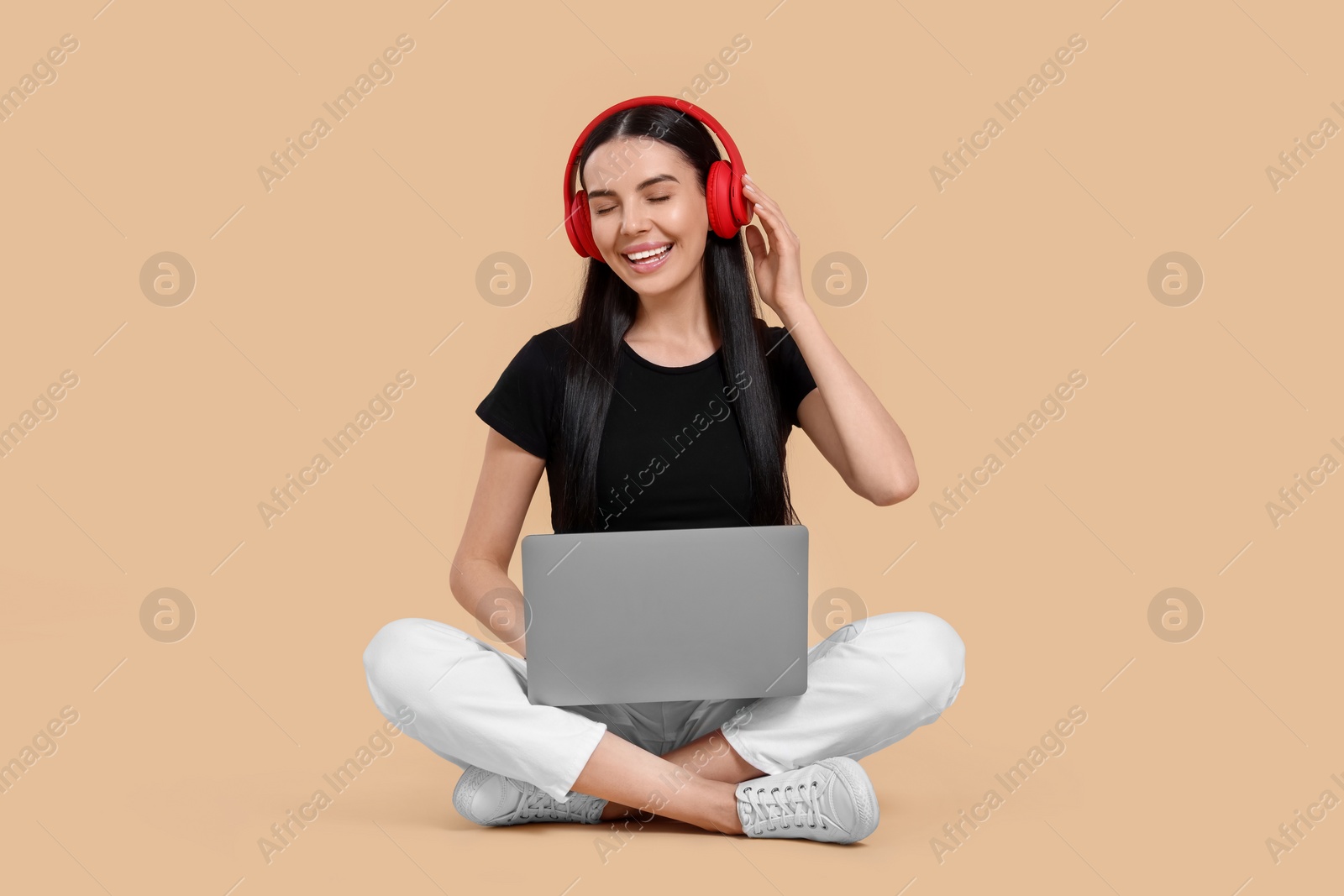 Photo of Happy woman with laptop listening to music in headphones on beige background