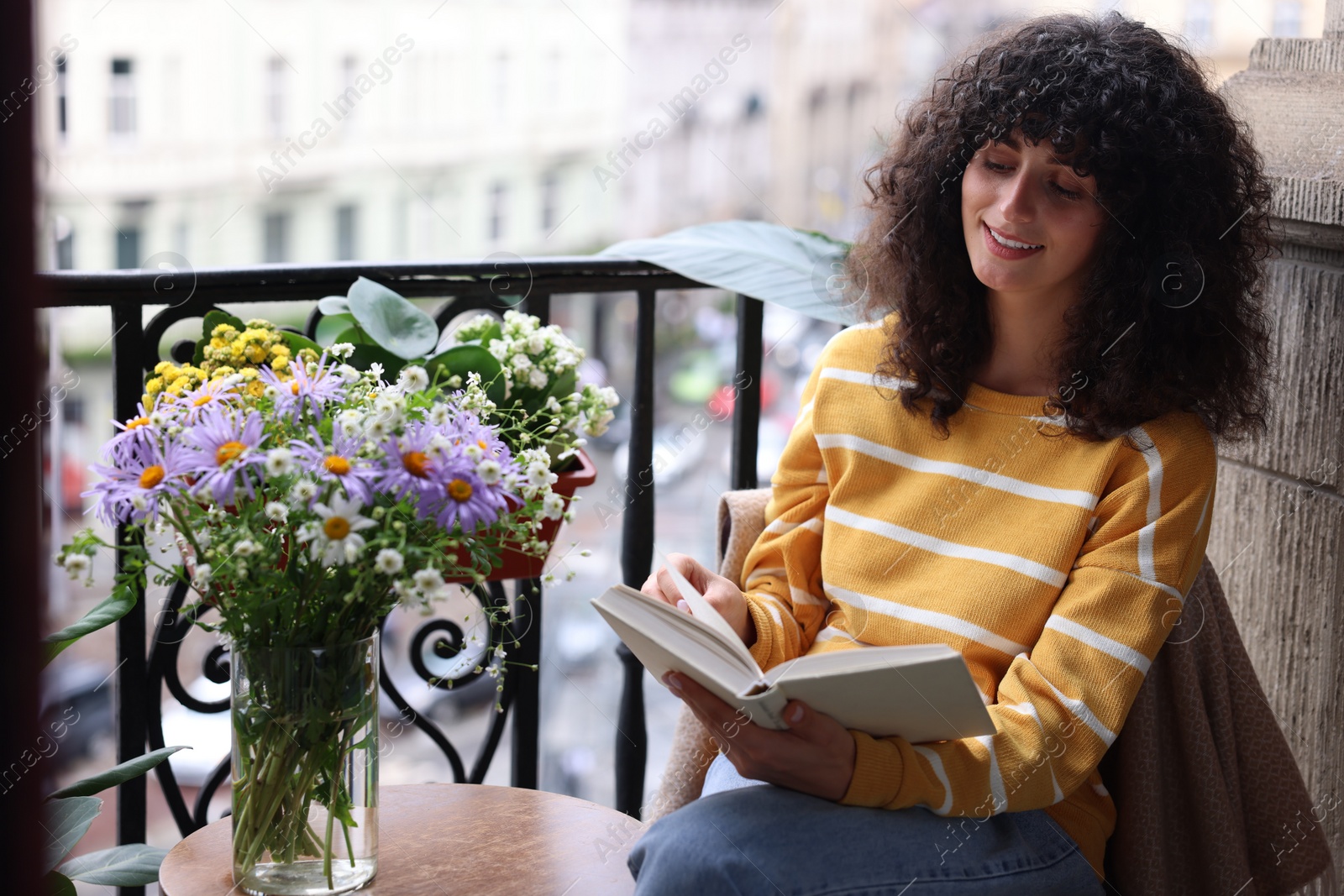 Photo of Young woman reading book at table with beautiful flowers on balcony