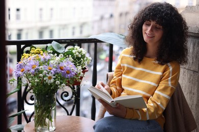 Photo of Young woman reading book at table with beautiful flowers on balcony