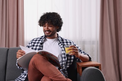 Photo of Happy man holding glass of juice and reading magazine on sofa with wooden armrest table at home