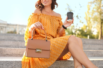 Photo of Young woman with stylish brown bag and cup of coffee on stairs outdoors, closeup