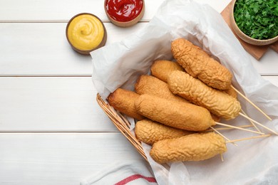 Photo of Delicious deep fried corn dogs in basket and sauces on white wooden table, flat lay. Space for text