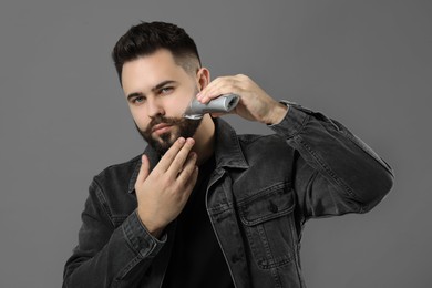 Handsome young man trimming beard on grey background