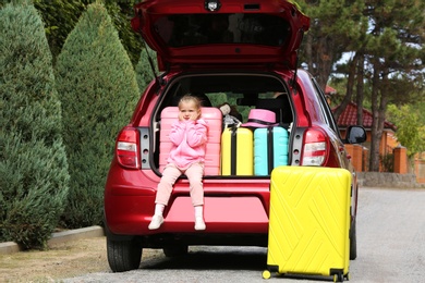 Photo of Cute little girl sitting in car trunk loaded with suitcases outdoors