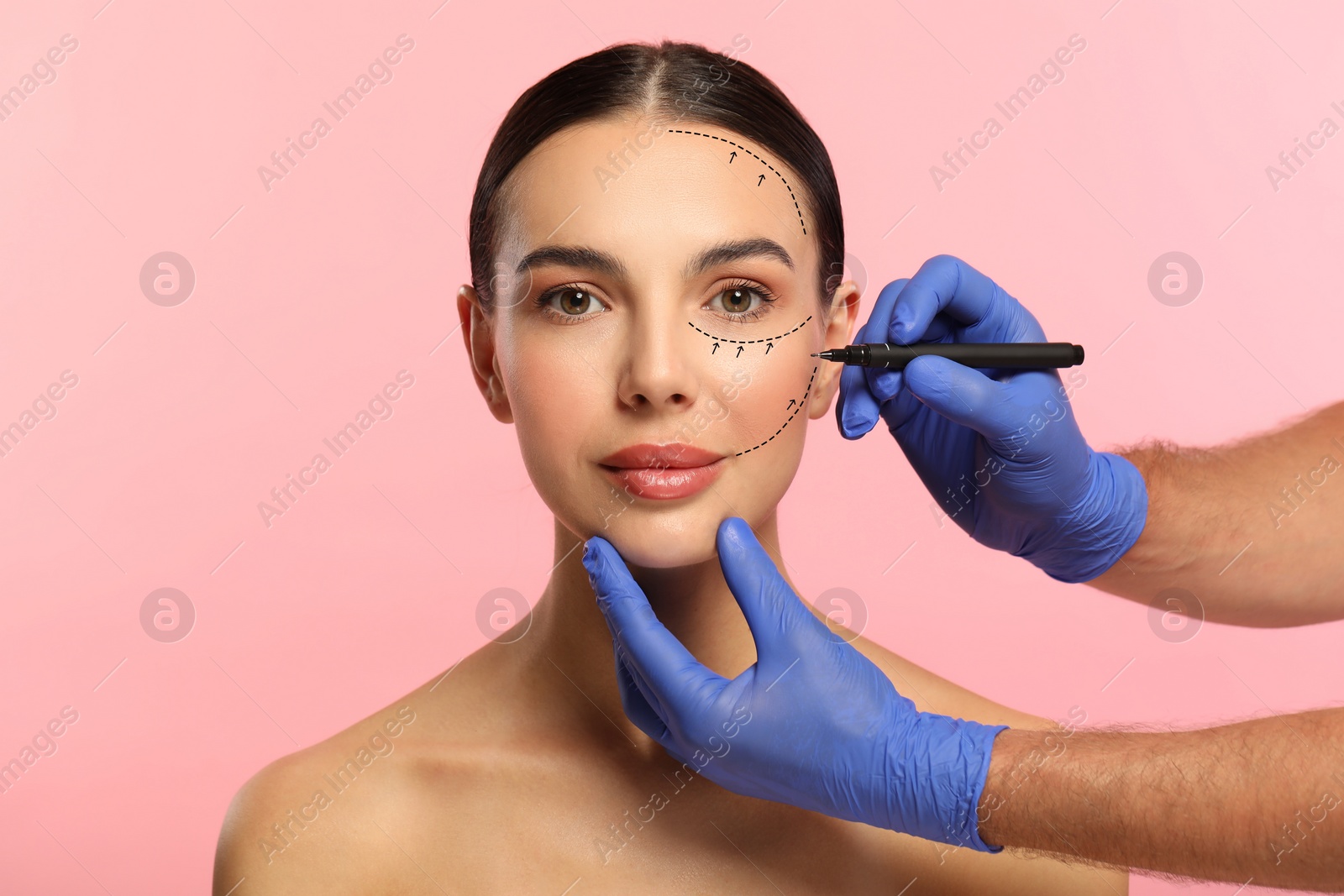 Image of Woman preparing for cosmetic surgery, pink background. Doctor drawing markings on her face, closeup