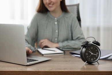 Photo of Black alarm clock and woman working on laptop at table indoors, closeup