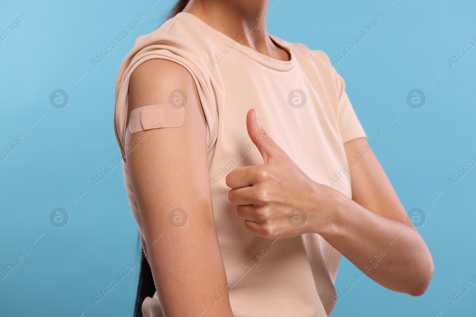 Photo of Woman with sticking plaster on arm after vaccination showing thumbs up against light blue background, closeup