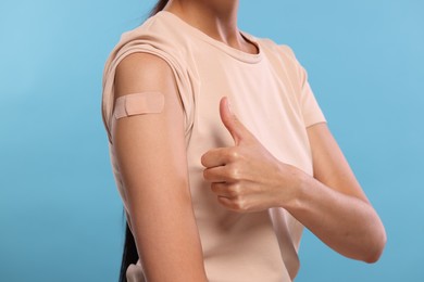 Photo of Woman with sticking plaster on arm after vaccination showing thumbs up against light blue background, closeup