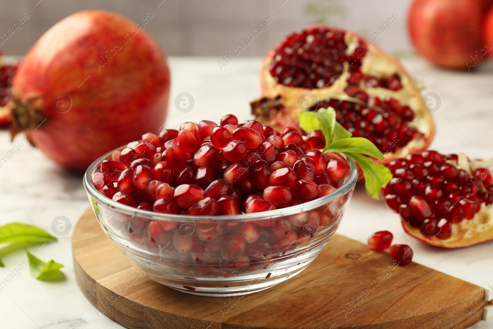 Photo of Ripe juicy pomegranate grains in bowl and green leaves on white table