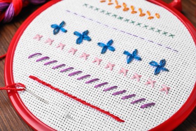 Fabric with colorful stitches and needle in embroidery hoop on wooden table, closeup