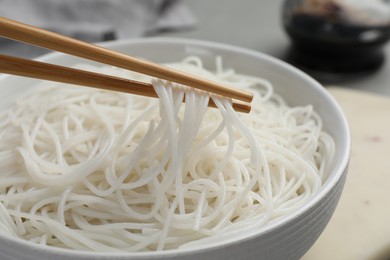 Photo of Chopsticks with cooked rice noodles over bowl on table, closeup