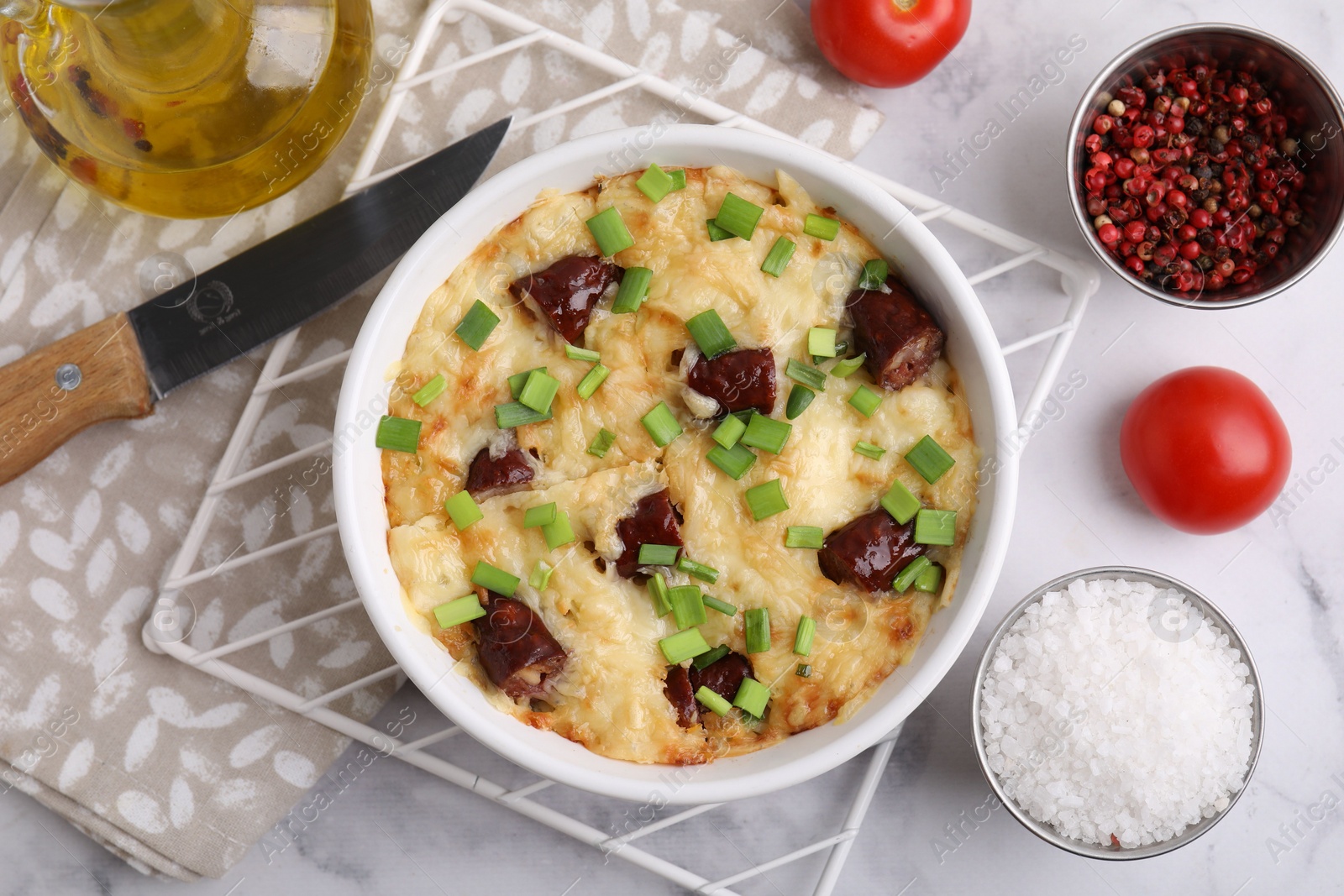 Photo of Tasty sausage casserole in baking dish, knife and ingredients on white marble table, flat lay