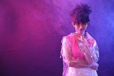 Photo of Beautiful young woman in transparent coat and sunglasses posing on color background in neon lights and smoke. Space for text