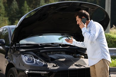 Photo of Stressed man talking on smartphone while looking under hood of broken car outdoors