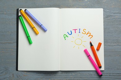 Photo of Colorful felt tip pens and notebook with word Autism on grey wooden table, top view