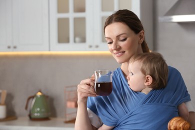 Photo of Mother with cup of drink holding her child in sling (baby carrier) in kitchen