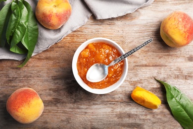 Flat lay composition with bowl of tasty peach jam, spoon and fresh fruit on wooden table