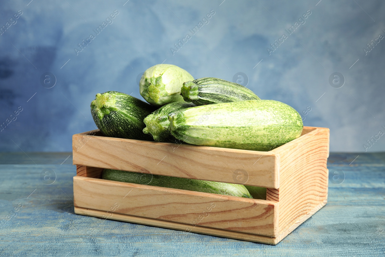 Photo of Wooden crate with fresh ripe green zucchinis on table against blue background