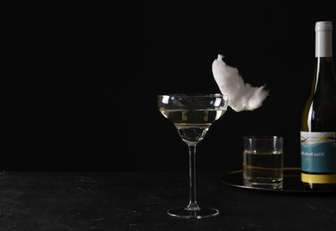 Photo of Cocktail with tasty cotton candy and bottle of alcohol drink on dark textured table against black background, space for text
