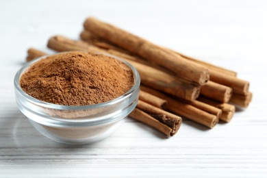 Photo of Aromatic cinnamon powder in bowl near sticks on white table, closeup