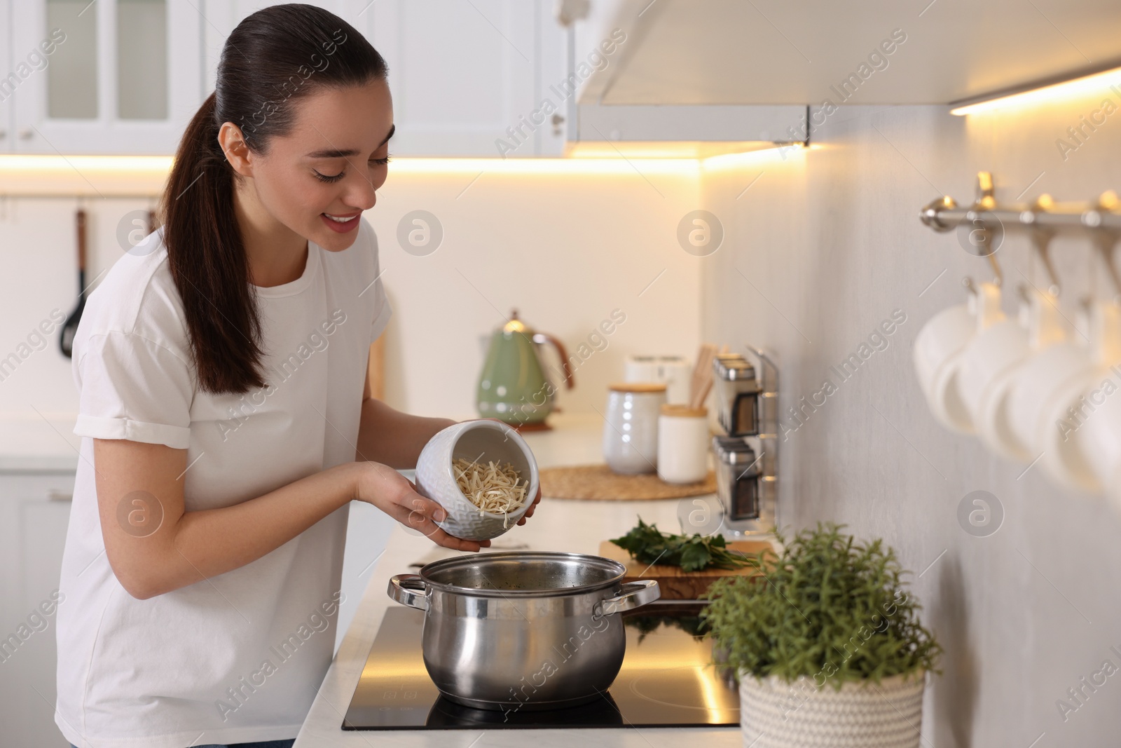 Photo of Smiling woman adding noodles into pot with soup in kitchen