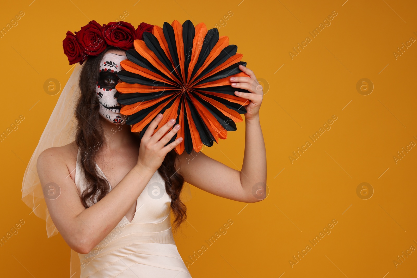 Photo of Young woman in scary bride costume with sugar skull makeup, flower crown and paper decoration on orange background, space for text. Halloween celebration