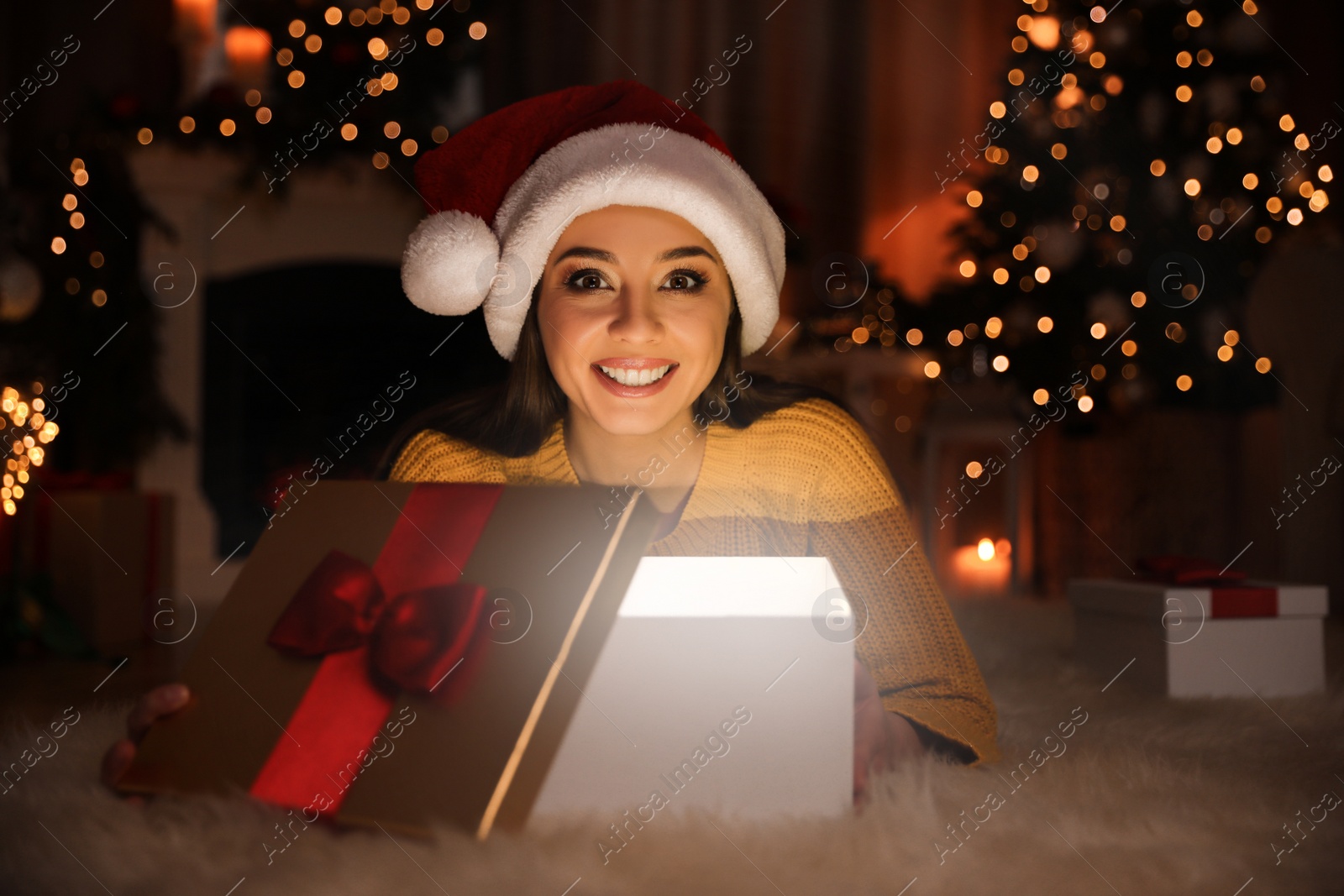 Photo of Young woman wearing Santa hat opening Christmas gift on floor at home