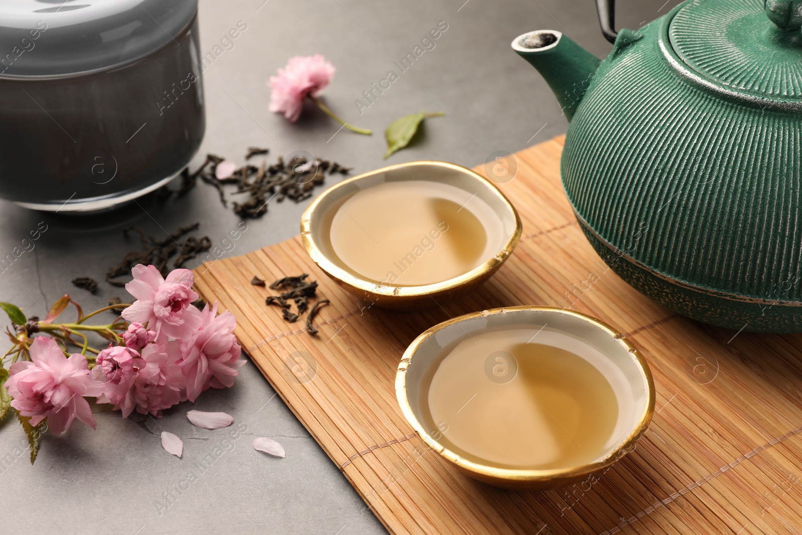 Photo of Traditional ceremony. Cups of brewed tea, teapot and sakura flowers on grey table, closeup