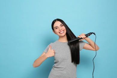 Photo of Beautiful happy woman showing thumbs up while using hair iron on light blue background. Space for text