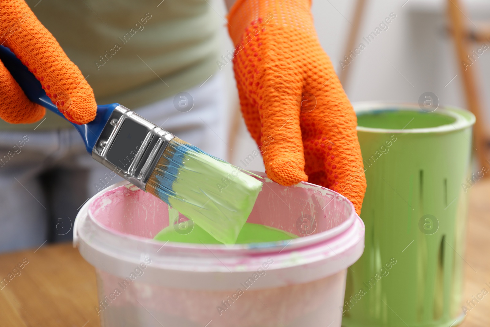 Photo of Woman dipping brush into bucket of green paint at table indoors, closeup