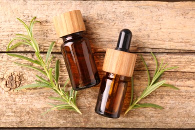 Aromatic essential oils in bottles and rosemary on wooden table, flat lay