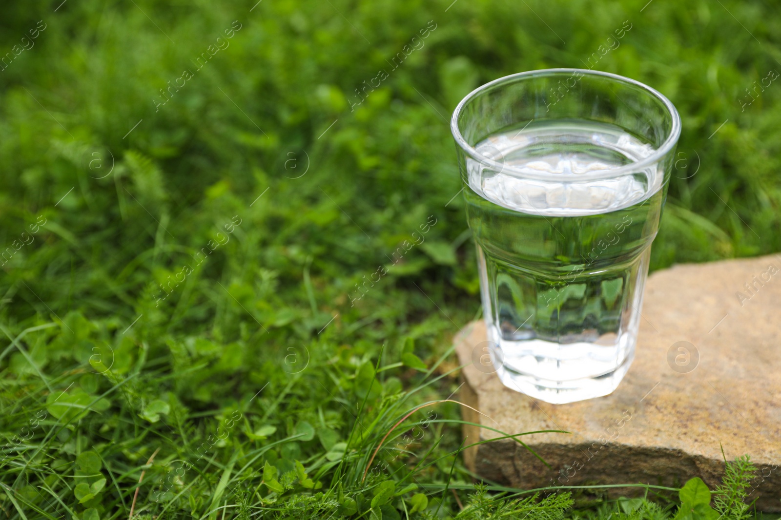 Photo of Glass of fresh water on stone in green grass outdoors. Space for text