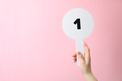 Woman holding auction paddle with number 1 on pink background, closeup. Space for text