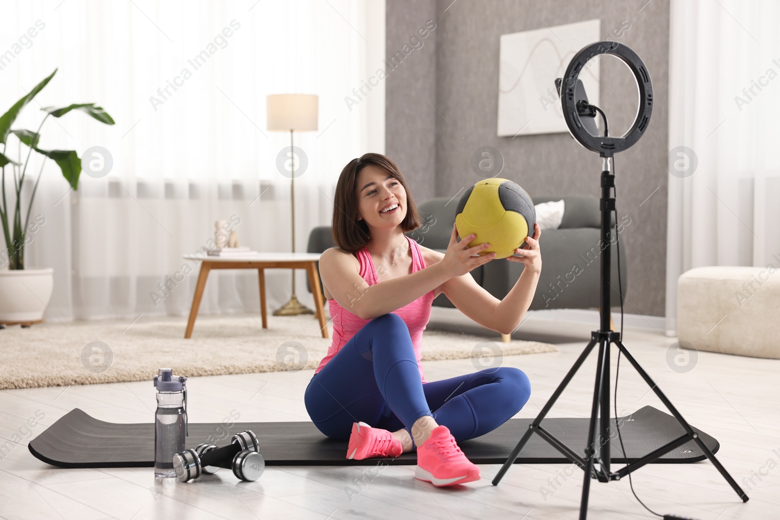 Photo of Happy sports blogger holding medicine ball while streaming online fitness lesson with smartphone at home