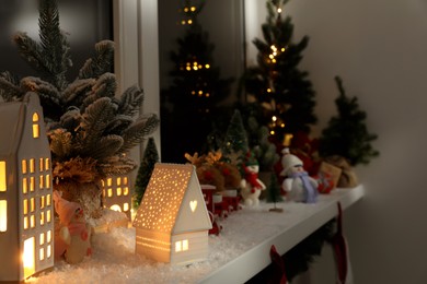 Photo of Christmas atmosphere. Beautiful glowing houses, fir trees, artificial snow and toys on window sill indoors