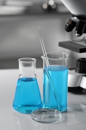 Photo of Different laboratory glassware with light blue liquid near microscope on table