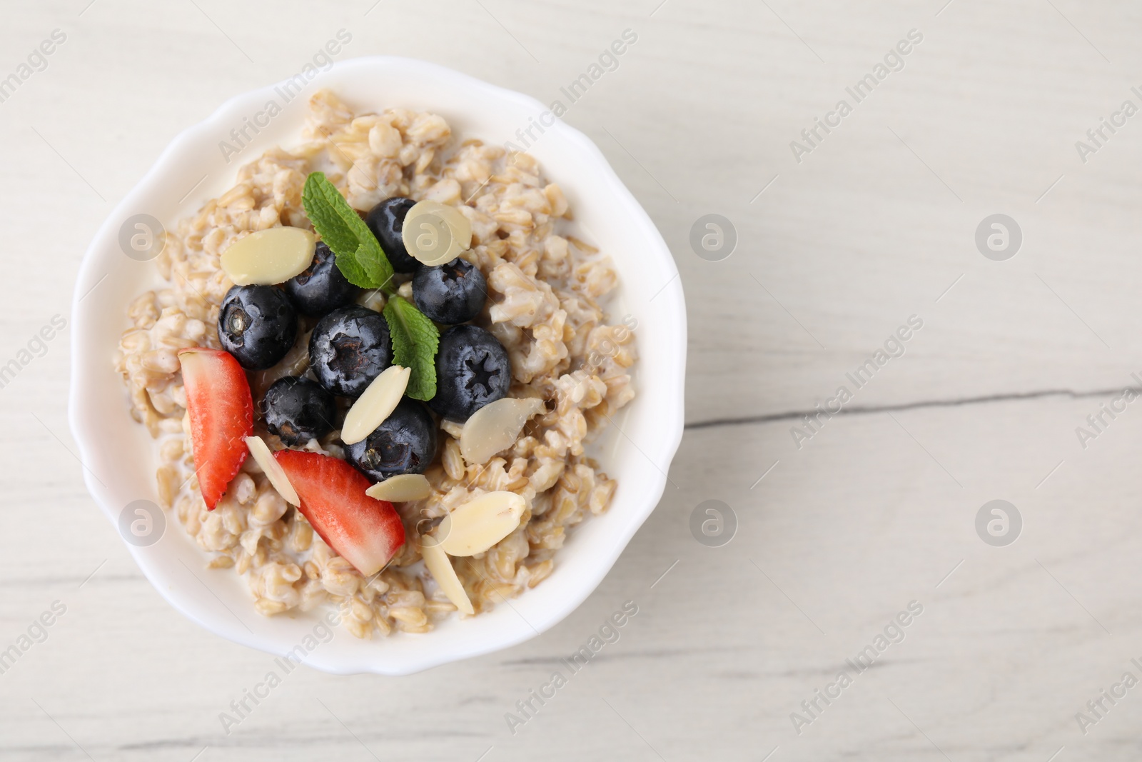 Photo of Tasty oatmeal with strawberries, blueberries and almond petals in bowl on white wooden table, top view. Space for text