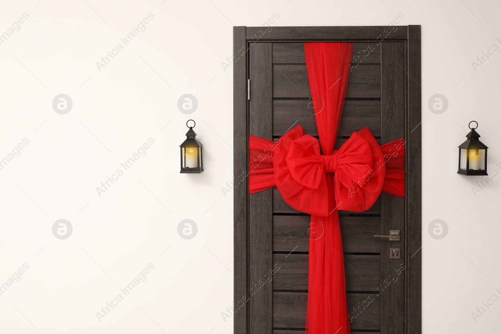 Photo of Wooden door with beautiful red bow and lanterns hanging on wall, space for text. Christmas decoration