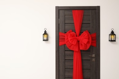 Photo of Wooden door with beautiful red bow and lanterns hanging on wall, space for text. Christmas decoration