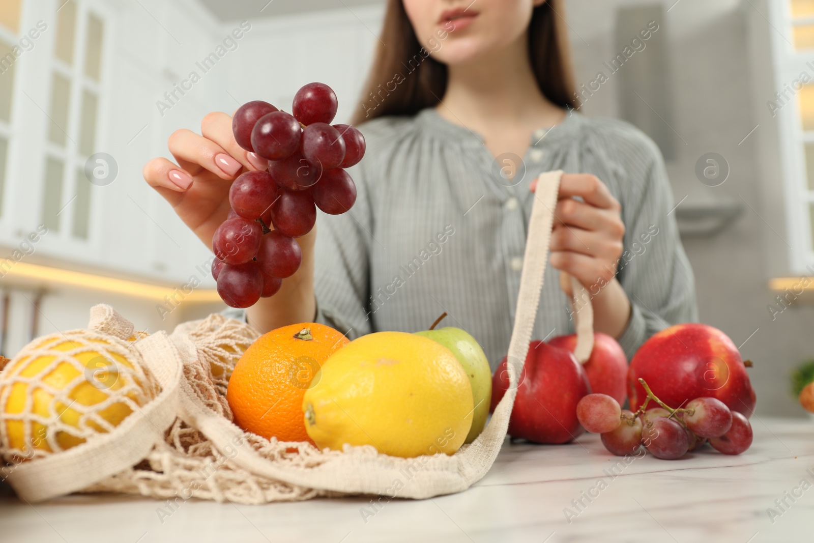 Photo of Woman taking grapes out from string bag at light marble table, closeup