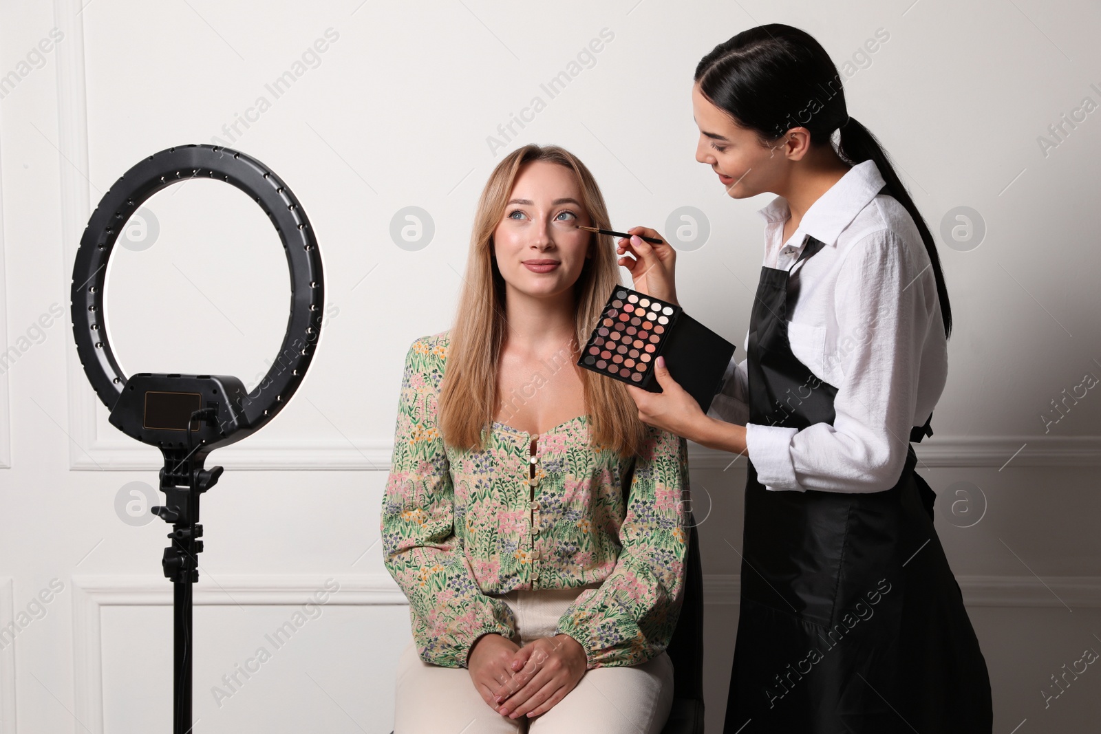 Photo of Professional makeup artist working with beautiful young woman near white wall. Using ring lamp