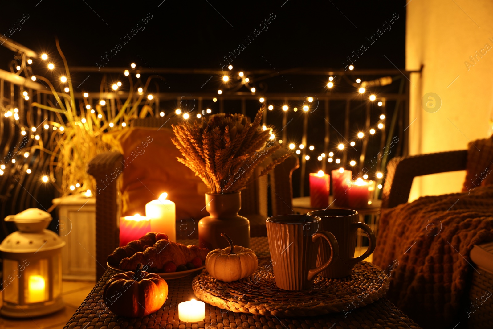 Photo of Rattan furniture, cups, fairy lights, burning candles and other autumn decor on outdoor terrace at night