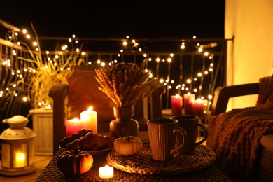 Rattan furniture, cups, fairy lights, burning candles and other autumn decor on outdoor terrace at night