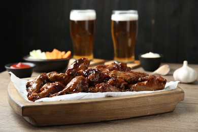 Photo of Tasty chicken wings, sauces, vegetable sticks and glasses of beer on wooden table, closeup. Delicious snacks