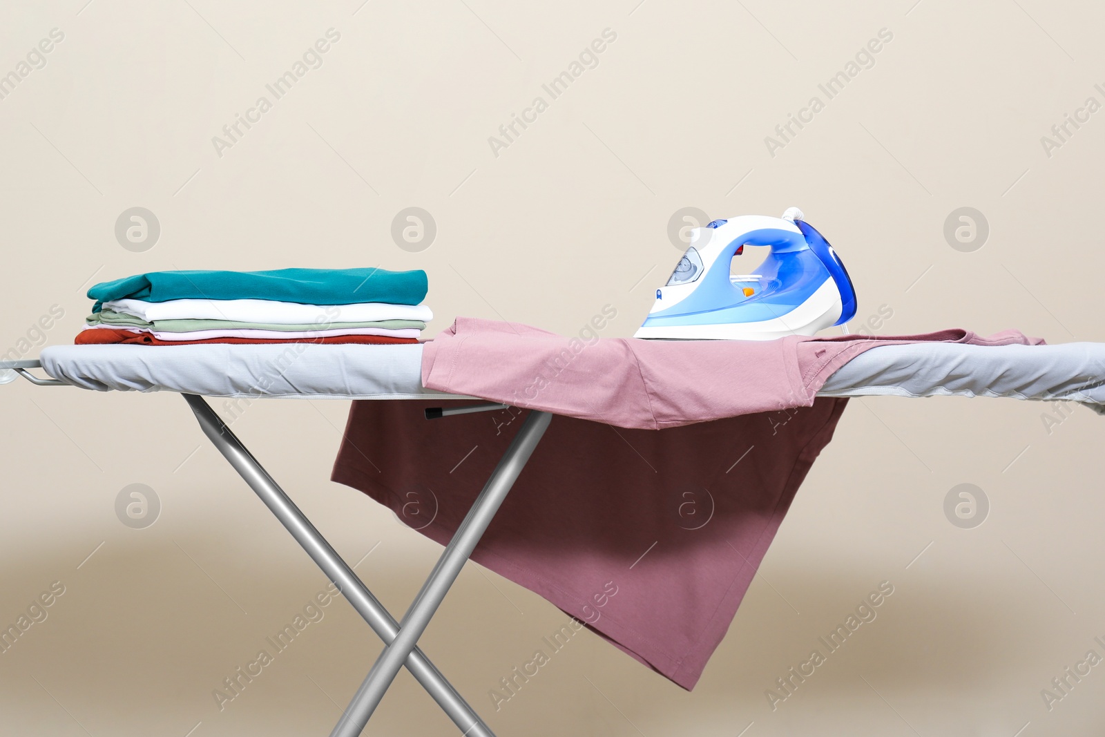 Photo of Modern iron and clean clothes on board near beige wall