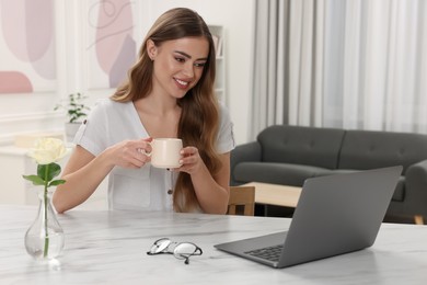 Photo of Happy woman with cup of drink using laptop at white table