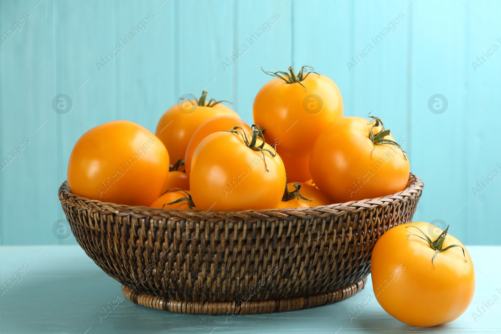 Photo of Ripe yellow tomatoes in wicker bowl on light blue wooden table