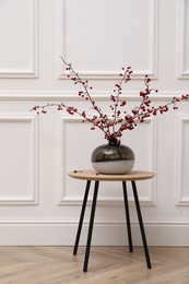 Photo of Hawthorn branches with red berries in vase on wooden table near white wall indoors