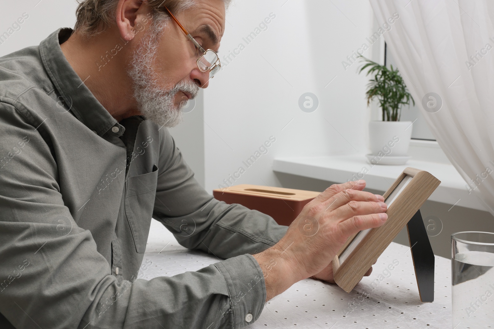 Photo of Upset senior man with photo frame at table in room. Loneliness concept